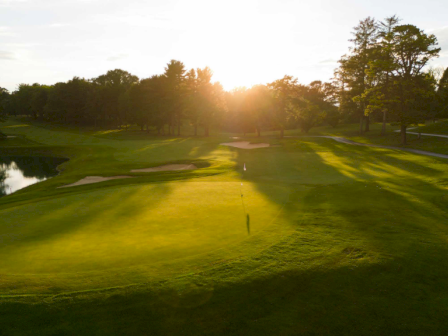 The image shows a serene golf course at sunset, featuring a green with sand traps, surrounded by trees and a small pond reflecting sunlight.