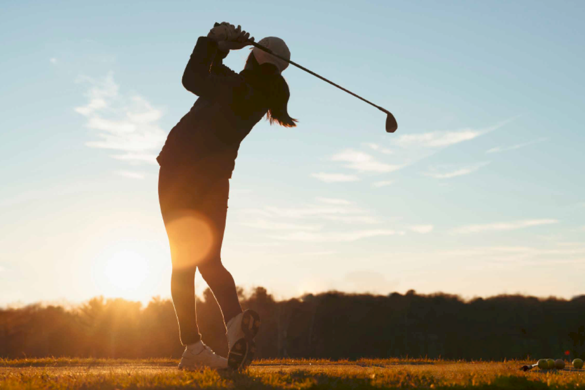 A golfer in mid-swing on a grassy field during sunset, with trees in the background.