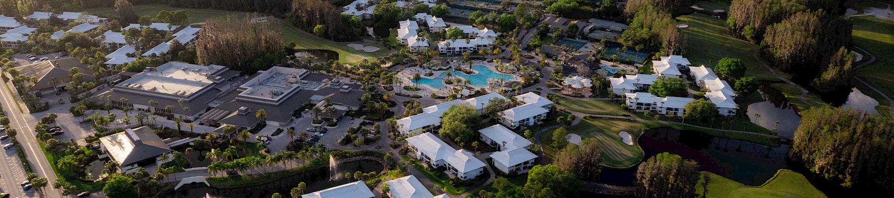 Aerial view of a resort with swimming pools, white-roofed buildings, tennis courts, and lush greenery.