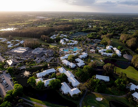 Aerial view of a resort with swimming pools, white-roofed buildings, tennis courts, and lush greenery.