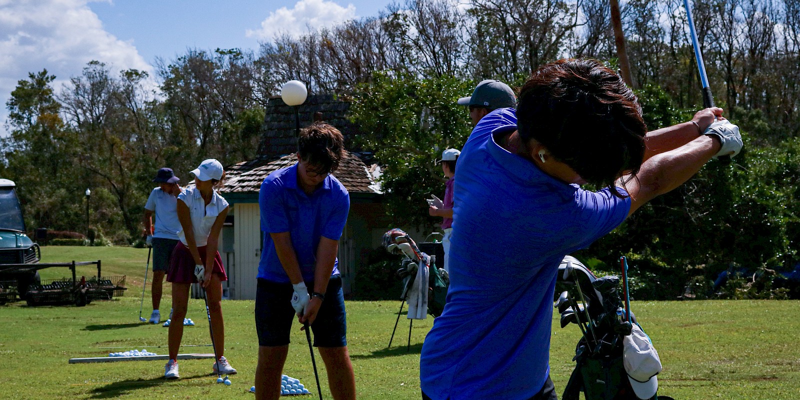 People practicing golf swings on a grassy field, with equipment bags nearby under a sunny sky.
