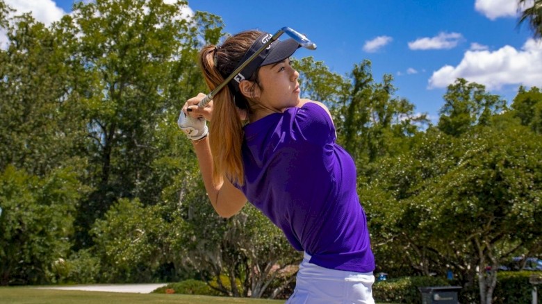 A person swings a golf club on a sunny day, wearing a purple shirt and white skirt, with trees and blue sky in the background.