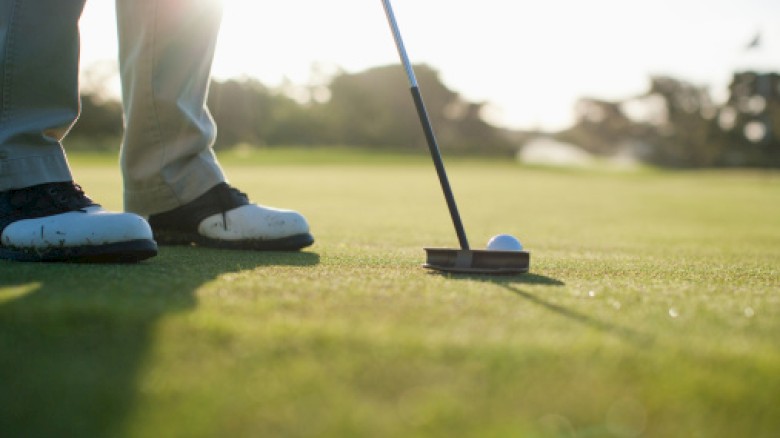 A person is on a golf course getting ready to putt a golf ball into the hole with a putter, under sunlight.
