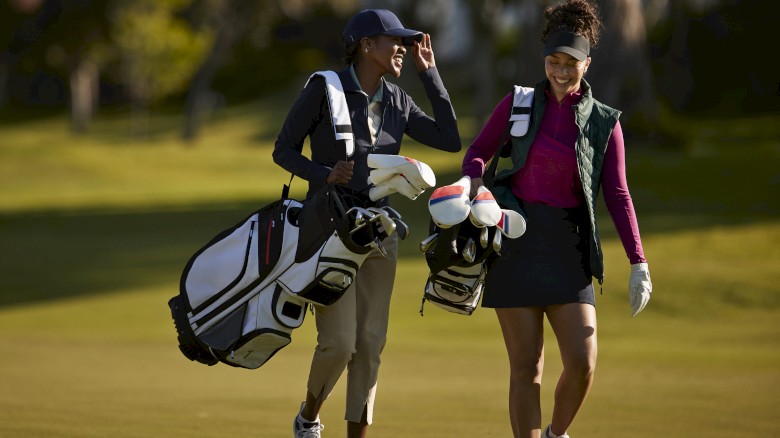 Two people are walking on a golf course, carrying golf bags and smiling, in a sunny outdoor setting.